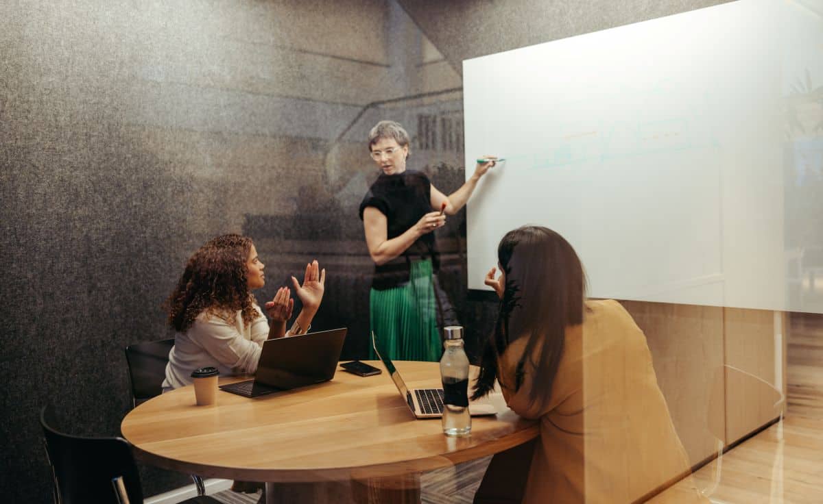 Une femme aux cheveux gris debout devant un tableau blanc parle à deux femmes assises à une table