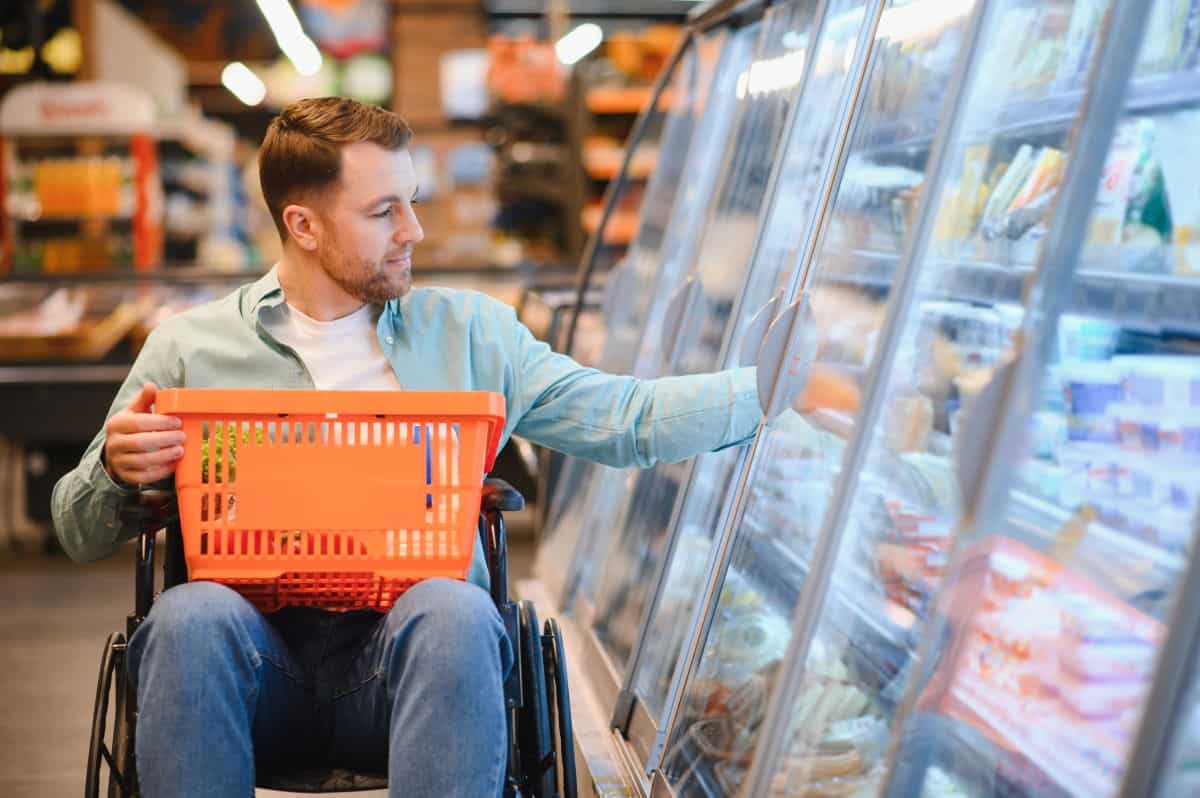 Un homme blanc en fauteuil roulant attrape un produit dans un meuble froid dans un supermarché