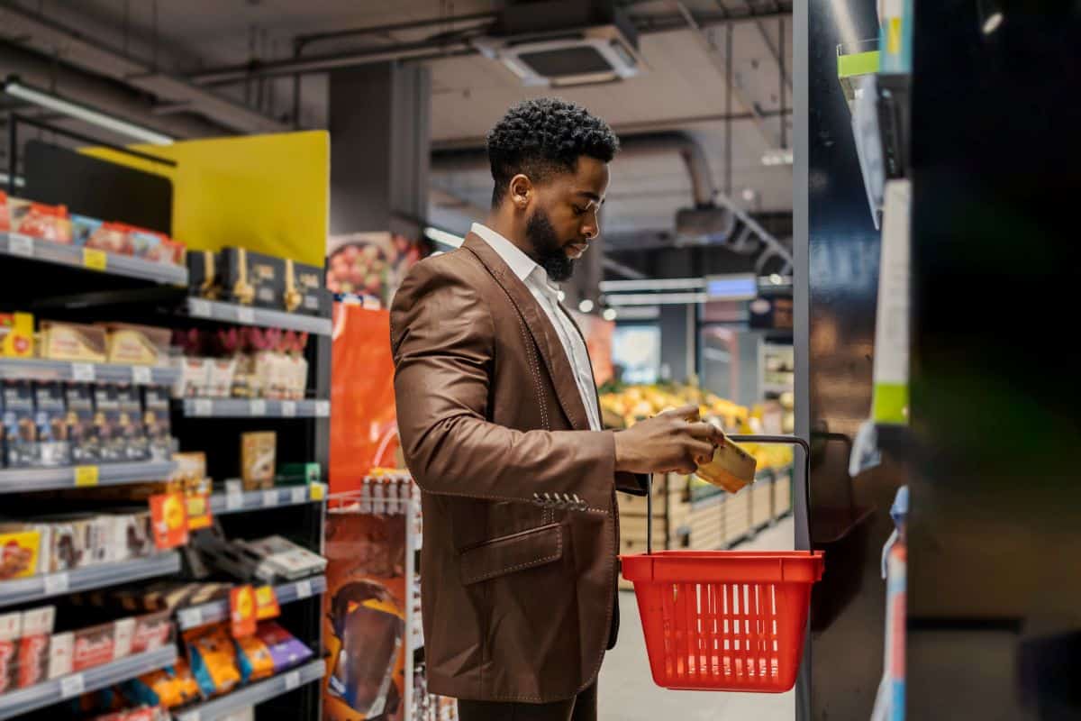 Un homme noir en costume marron dans un supermarché saisit un produit pour le poser dans son panier