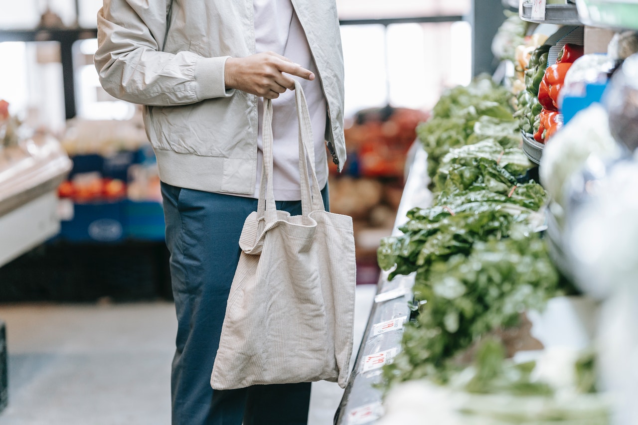 Une personne portant un jean, une veste grise et un sac en tissu à la main devant des légumes