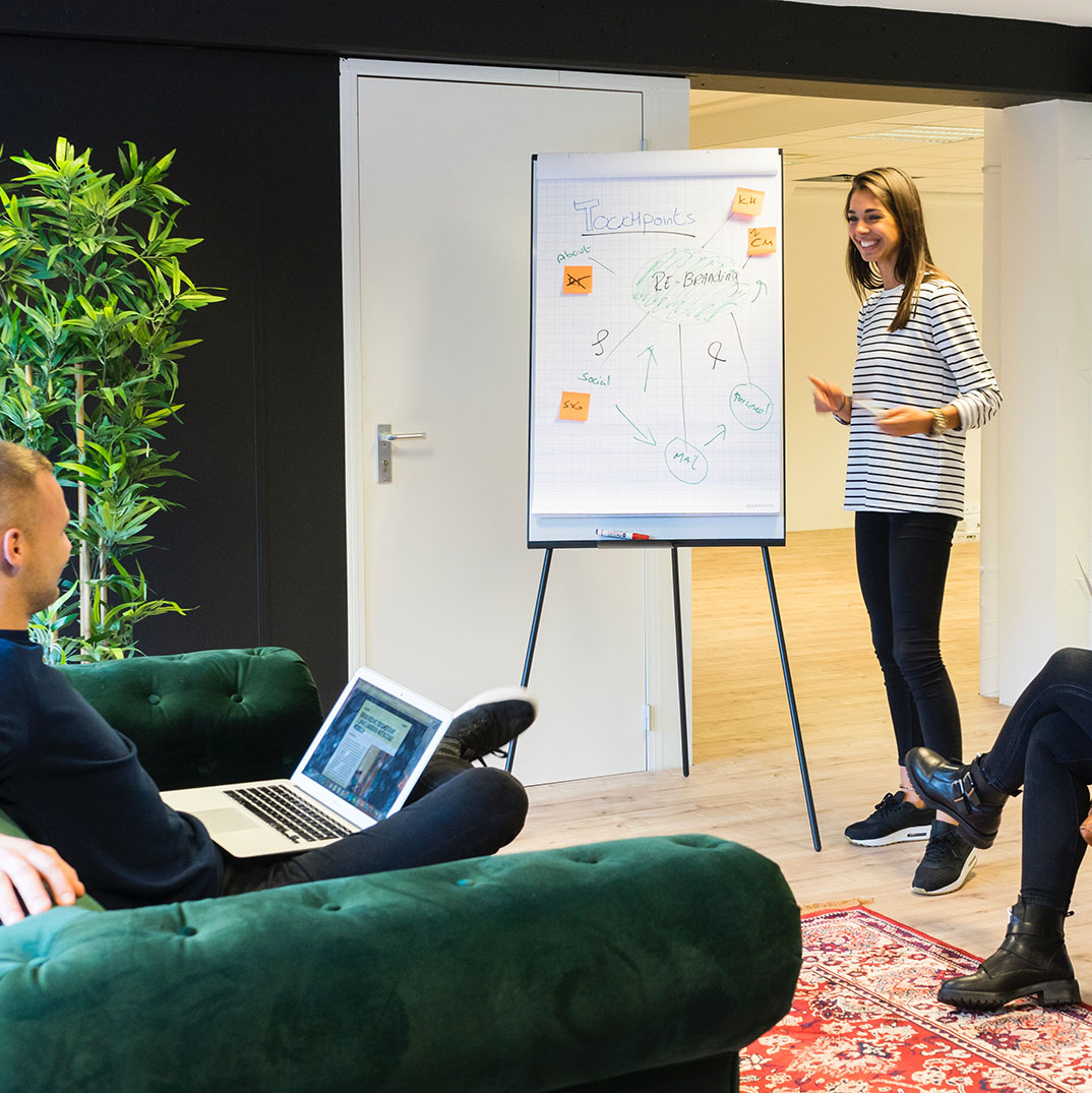 A gauche un homme blanc aux cheveux blonds courts assis dans un canapé vert face à une femme blanche debout devant un tableau blanc