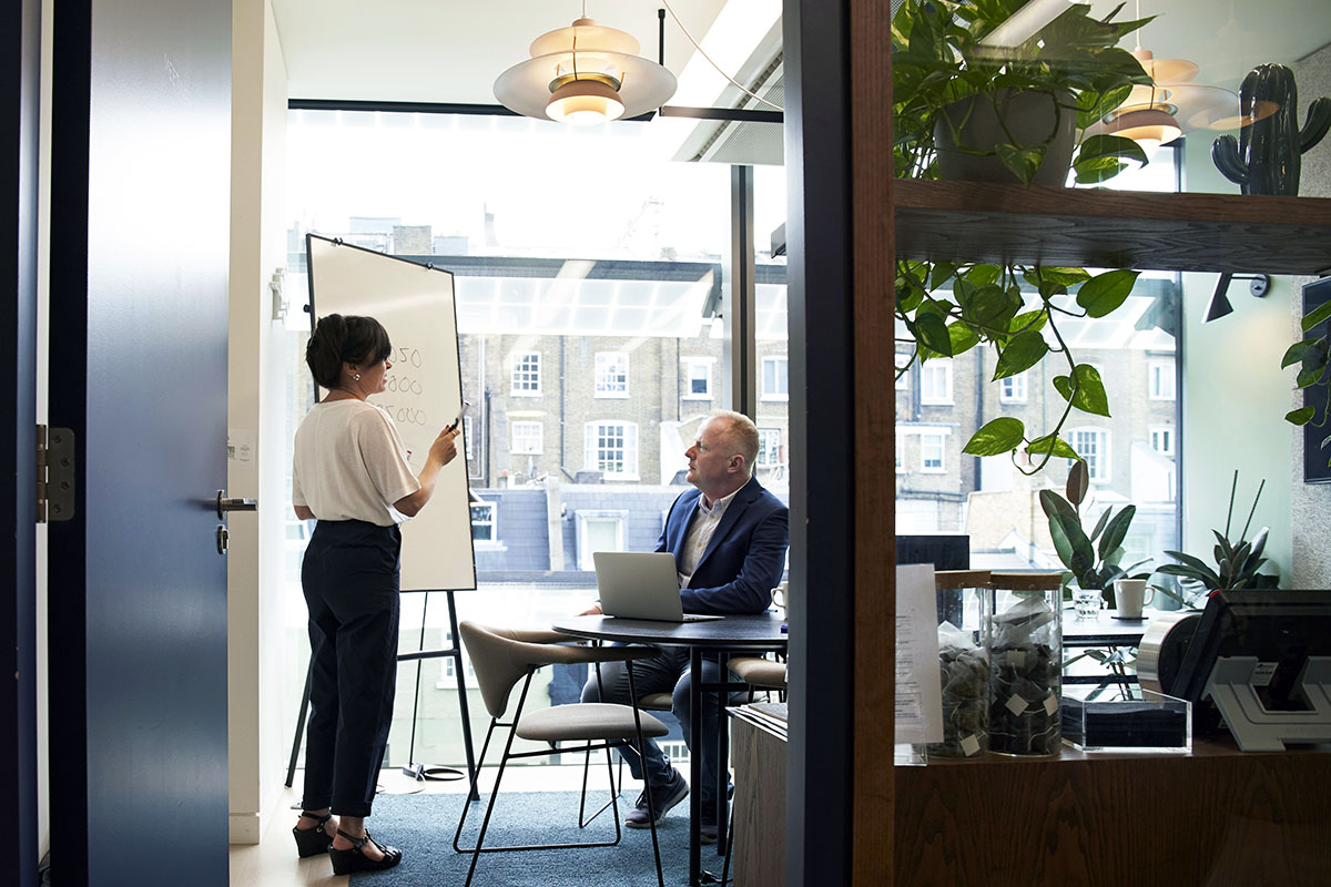 Dans un bureau une femme debout à la peau mate présente un tableau à un homme blanc