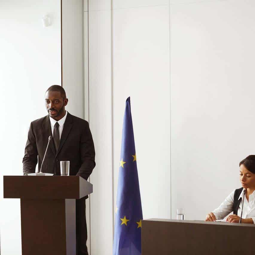 Un homme noir parle à un pupitre devant un drapeau européen à côté d'une femme à la peau mate qui écrit