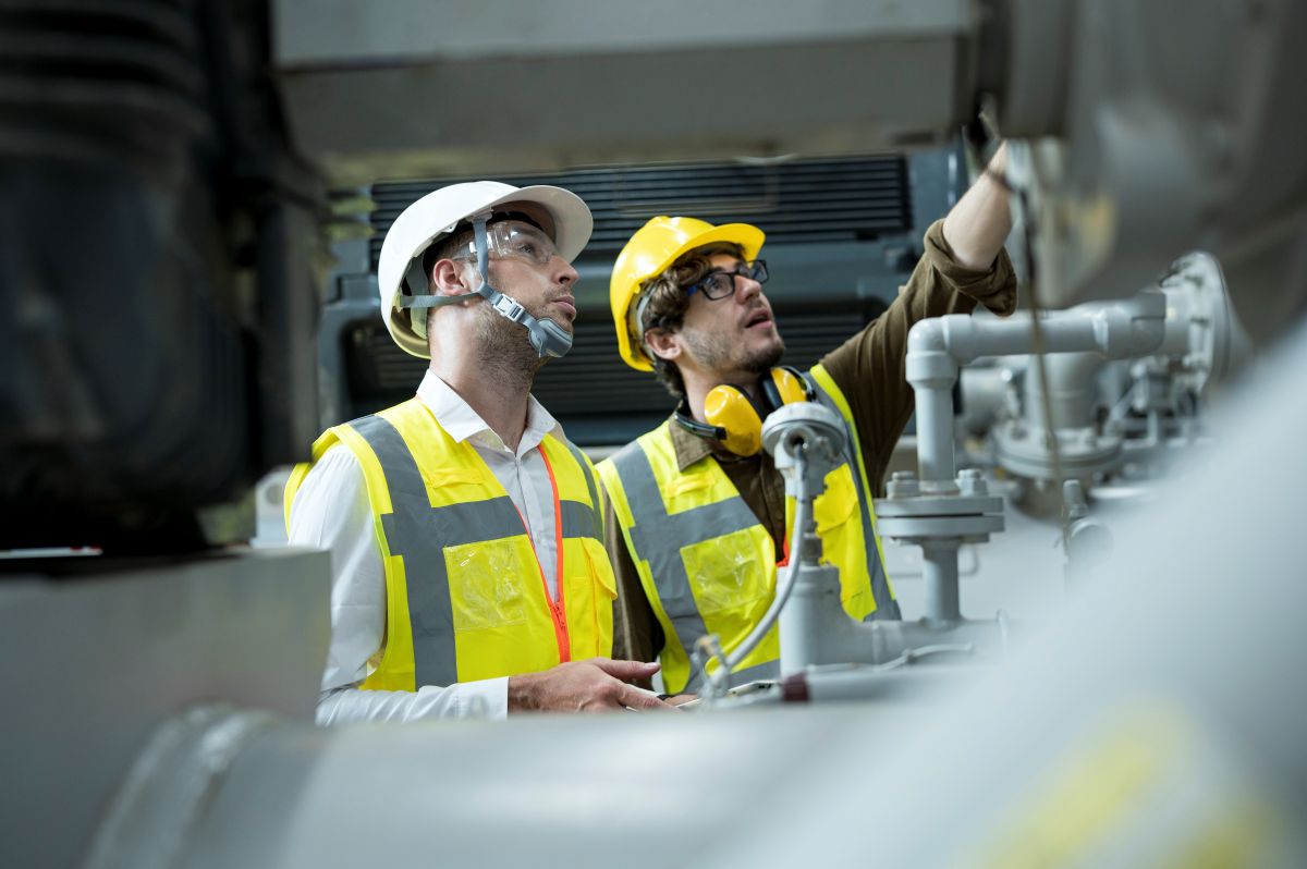 Deux hommes blancs en gilet jaune à côté de tuyaux dans une usine