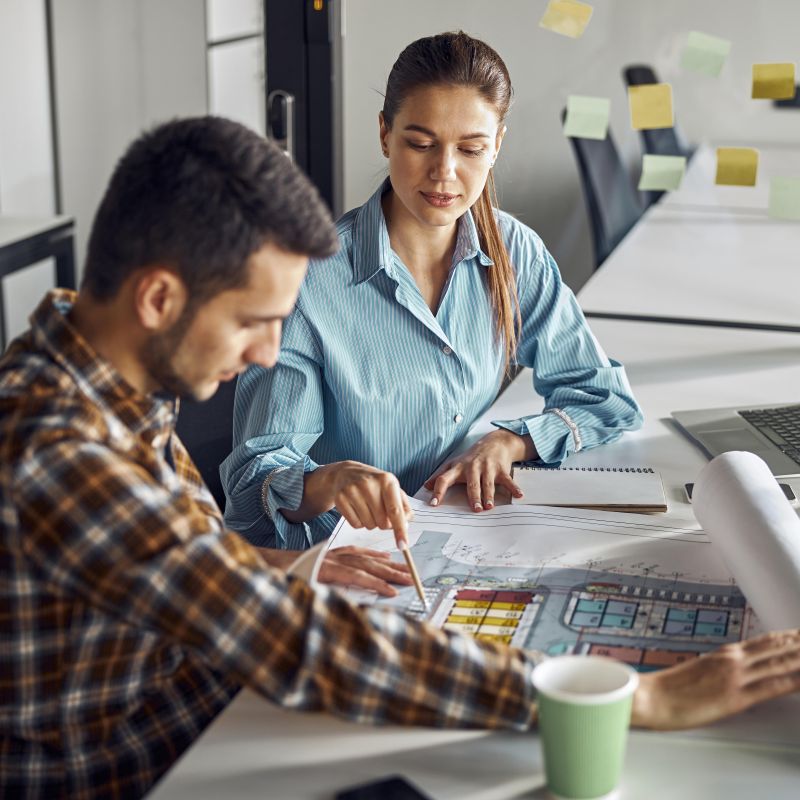 Une femme blanche pointe un plan du doigt à son collègue blanc assis à sa gauche